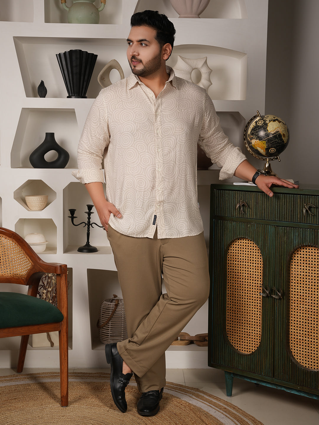 Man in a beige shirt and brown pants standing in a room with decorative shelves and furniture.